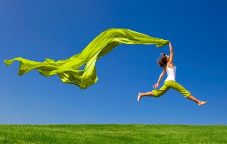 Frau mit Energie springt mit einem grünen Tuch über einen Rasen Blauer Himmel